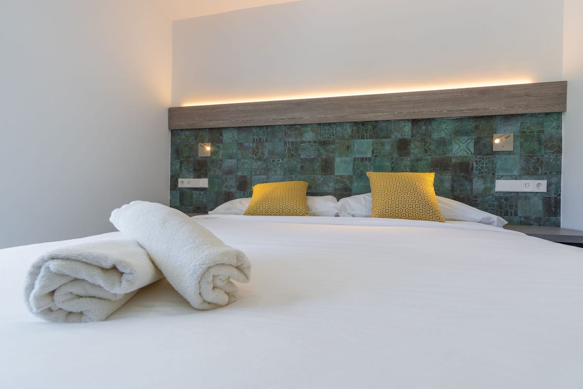 Una cama bien hecha con sábanas blancas y dos almohadas estampadas, un cabecero de madera y una pared con diseño de mosaico verde.