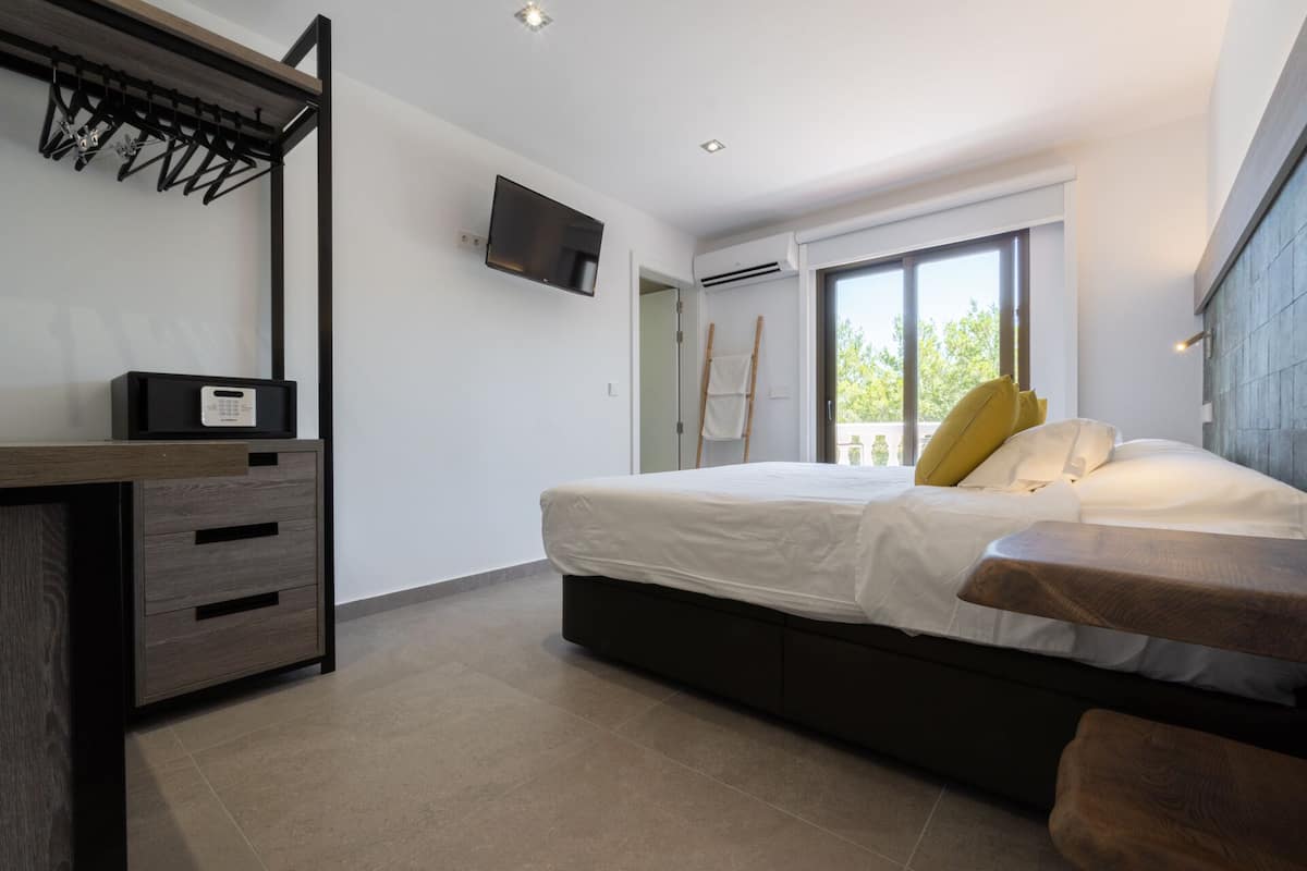 Un dormitorio moderno con cama, televisor montado en la pared, escritorio de madera y ventana con vista a zonas verdes.