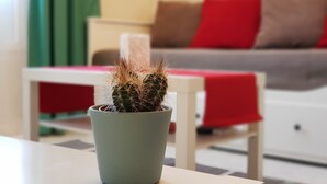 Um cacto num vaso sobre uma mesa numa sala de estar com um sofá e um cobertor vermelho ao fundo.