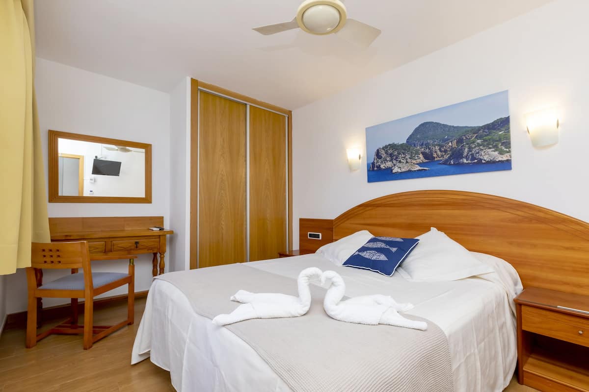 A hotel room with a bed, wooden headboard, a desk with a chair, a wardrobe, a painting of a coastal landscape, and a ceiling fan.