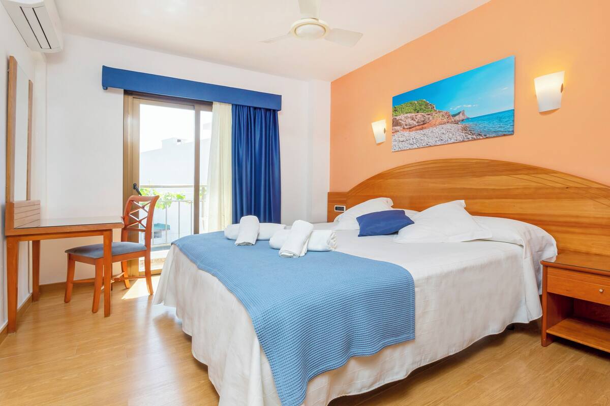 A hotel room with a bed, a desk, a chair, a window with curtains, a painting of a beach, and a ceiling fan.