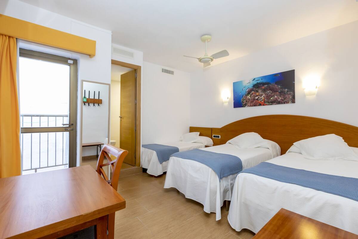 A hotel room with two beds, a wooden desk, a chair, a ceiling fan, and a wall-mounted picture.