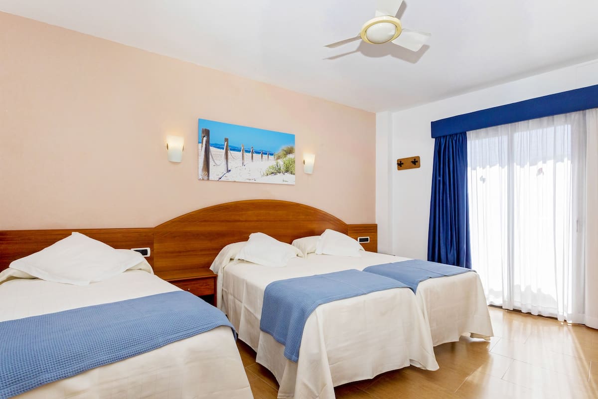 A hotel room with two beds, a ceiling fan, a painting of a beach, and a large window with blue curtains.