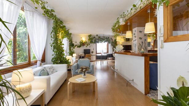 Amplia sala de estar con sofá, mesa de centro y mesa de comedor. Hay plantas colgando del techo y una cocina al fondo.