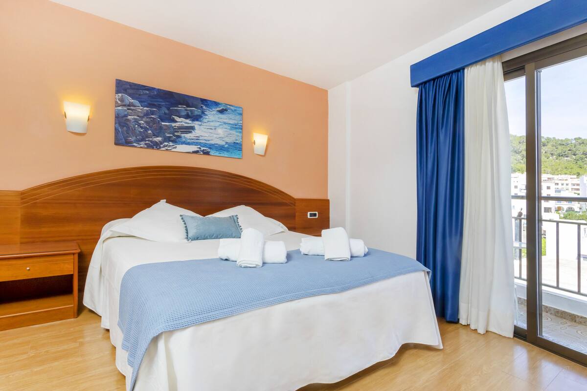 A hotel room with a wooden headboard, a bed with white and blue bedding, two wall-mounted lights, a painting, and a view of buildings through a large window with blue curtains.