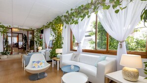 Una sala de estar moderna con un sofá blanco, un sillón azul y una pequeña mesa redonda. Hay cortinas blancas y plantas verdes colgando del techo.