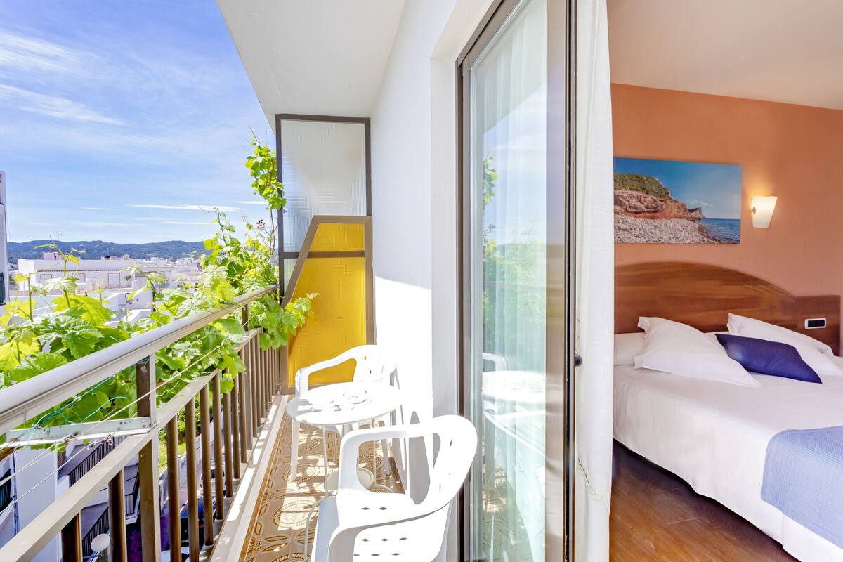 A balcony with white chairs and a yellow chair, a bed with white bedding and a blue pillow, and a painting of a beach on the wall.