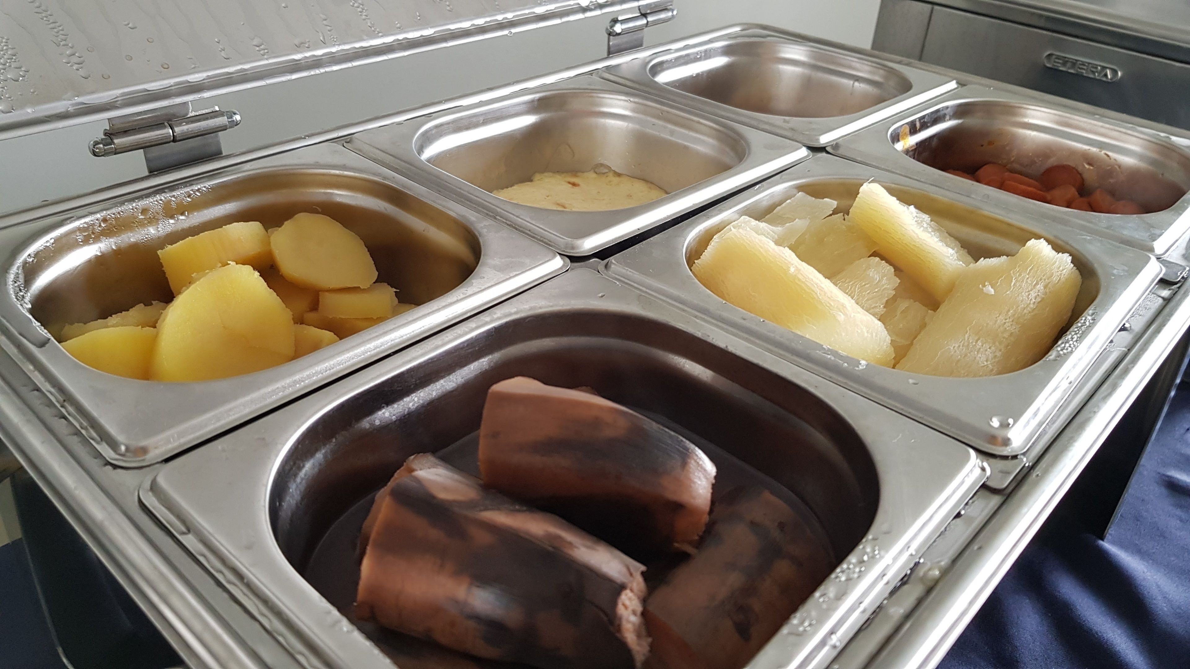 Un bufé con diversos alimentos, entre ellos patatas, salchichas y otros platos, presentados en recipientes de acero inoxidable.