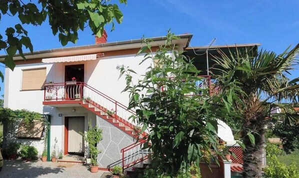 Uma casa de dois andares com varanda, escadaria e plantas em vasos.