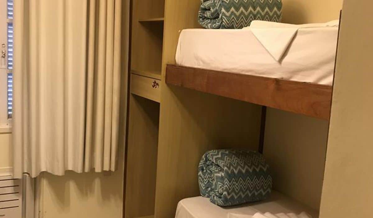 A bunk bed with a white mattress and a patterned pillow, a wooden shelf, and a white radiator.