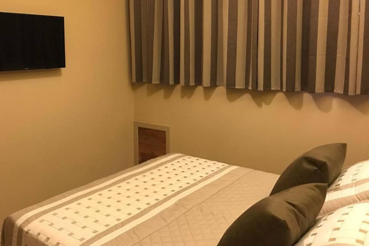 Une chambre d’hôtel équipée d’un lit, d’un téléviseur à écran plat fixé au mur et d’une fenêtre avec des stores verticaux.