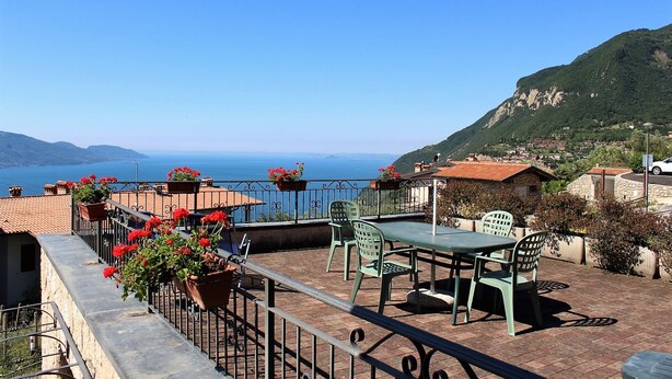 A terrace with a table and chairs, overlooking a coastal landscape with a view of the sea and mountains.