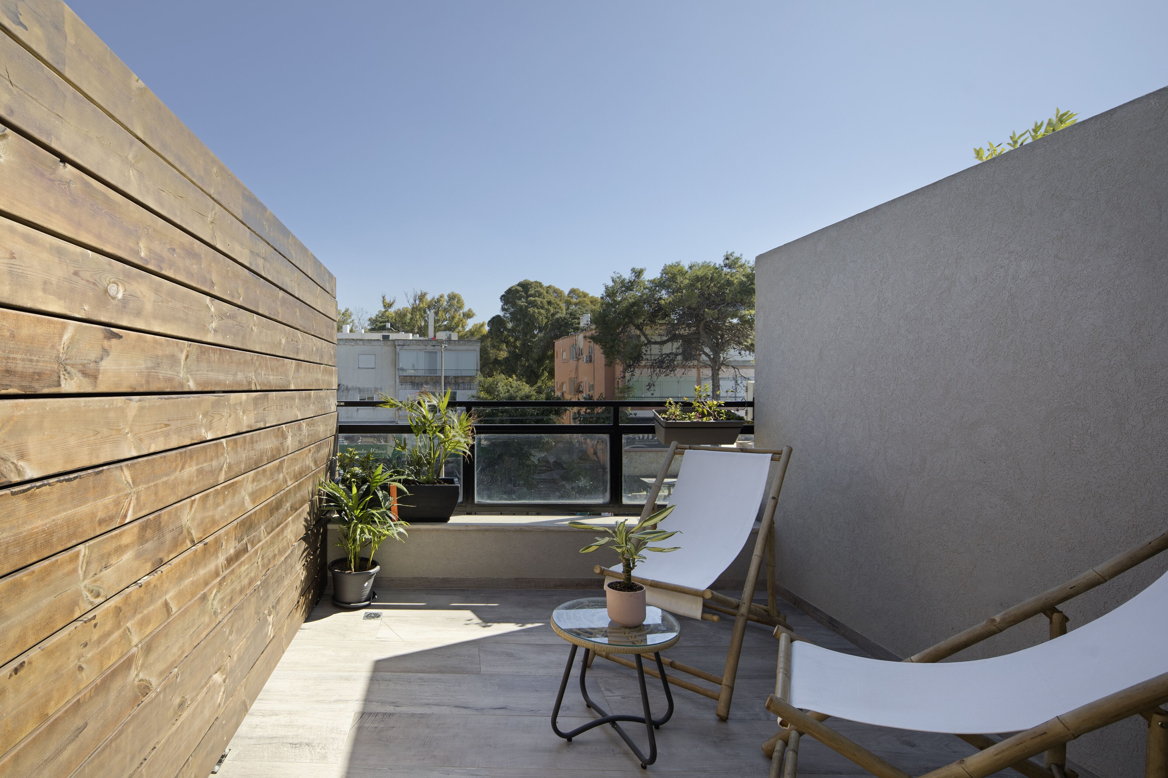 Un balcon avec un mur en bois, une rambarde en verre, une chaise blanche, une petite table sur laquelle est posée une plante en pot, et une vue sur des immeubles et des arbres.