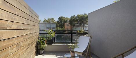 Un balcon avec un mur en bois, une rambarde en verre, une chaise blanche, une petite table sur laquelle est posée une plante en pot, et une vue sur des immeubles et des arbres.