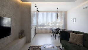 Uma sala de estar moderna com uma televisão de tela plana, um sofá verde, uma área de jantar com mesa e cadeiras, e uma janela grande com cortinas leves.