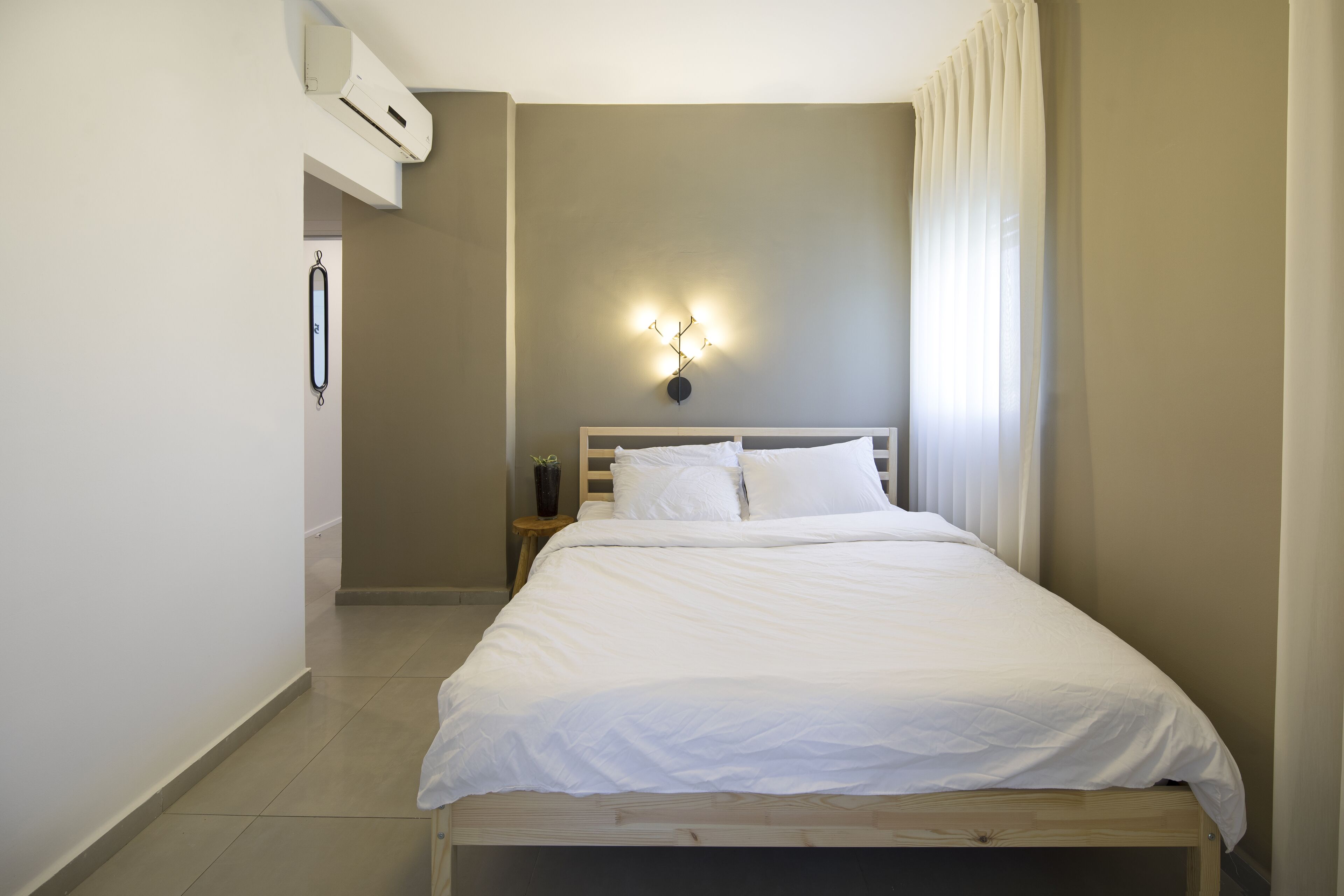 Un letto ben rifatto in una camera d'albergo, con una testiera, un condizionatore d'aria a muro e un comodino con una lampada.