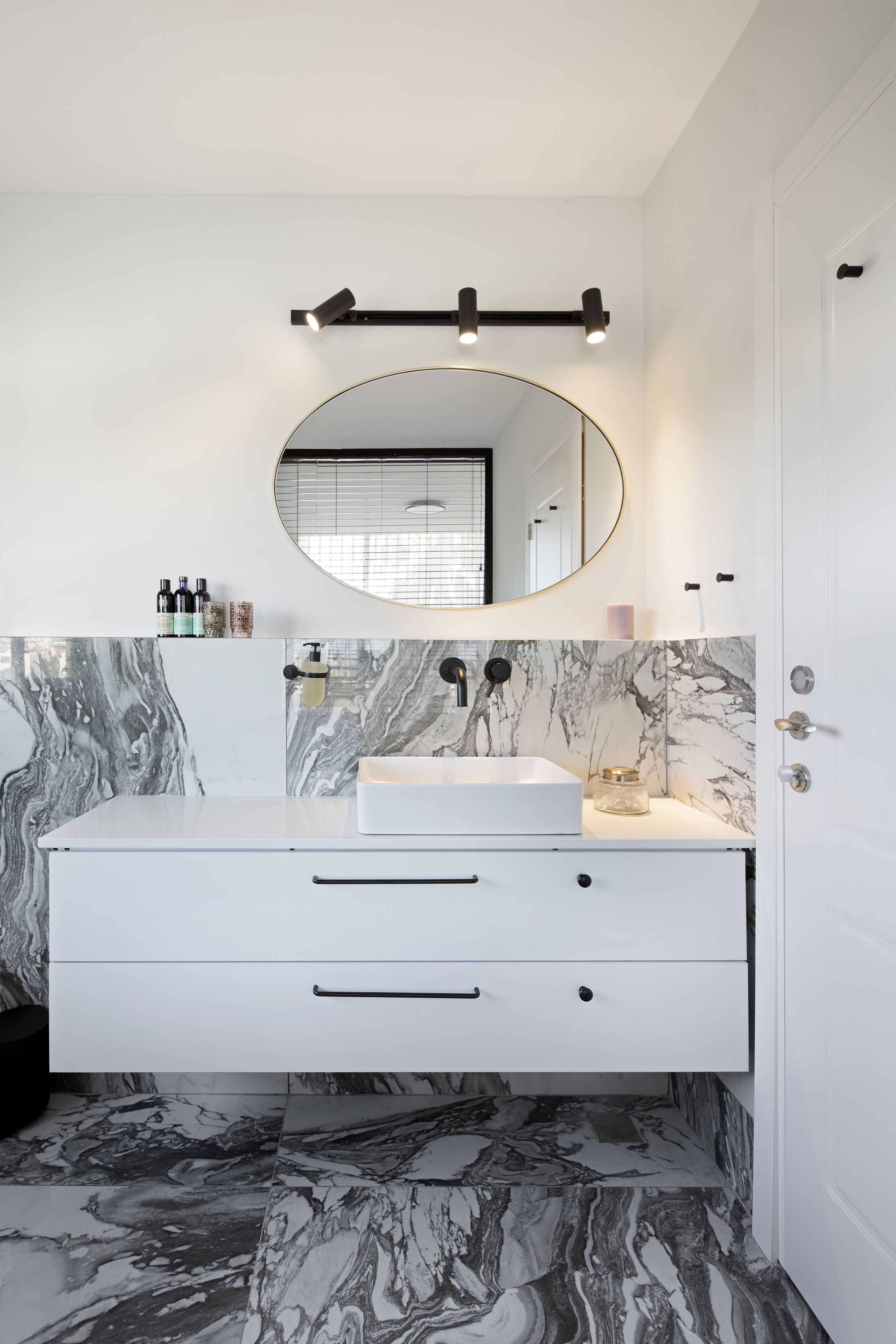Een badkamer met een witte wastafelmeubel, een marmeren spatwand en een ronde spiegel boven de wastafel.