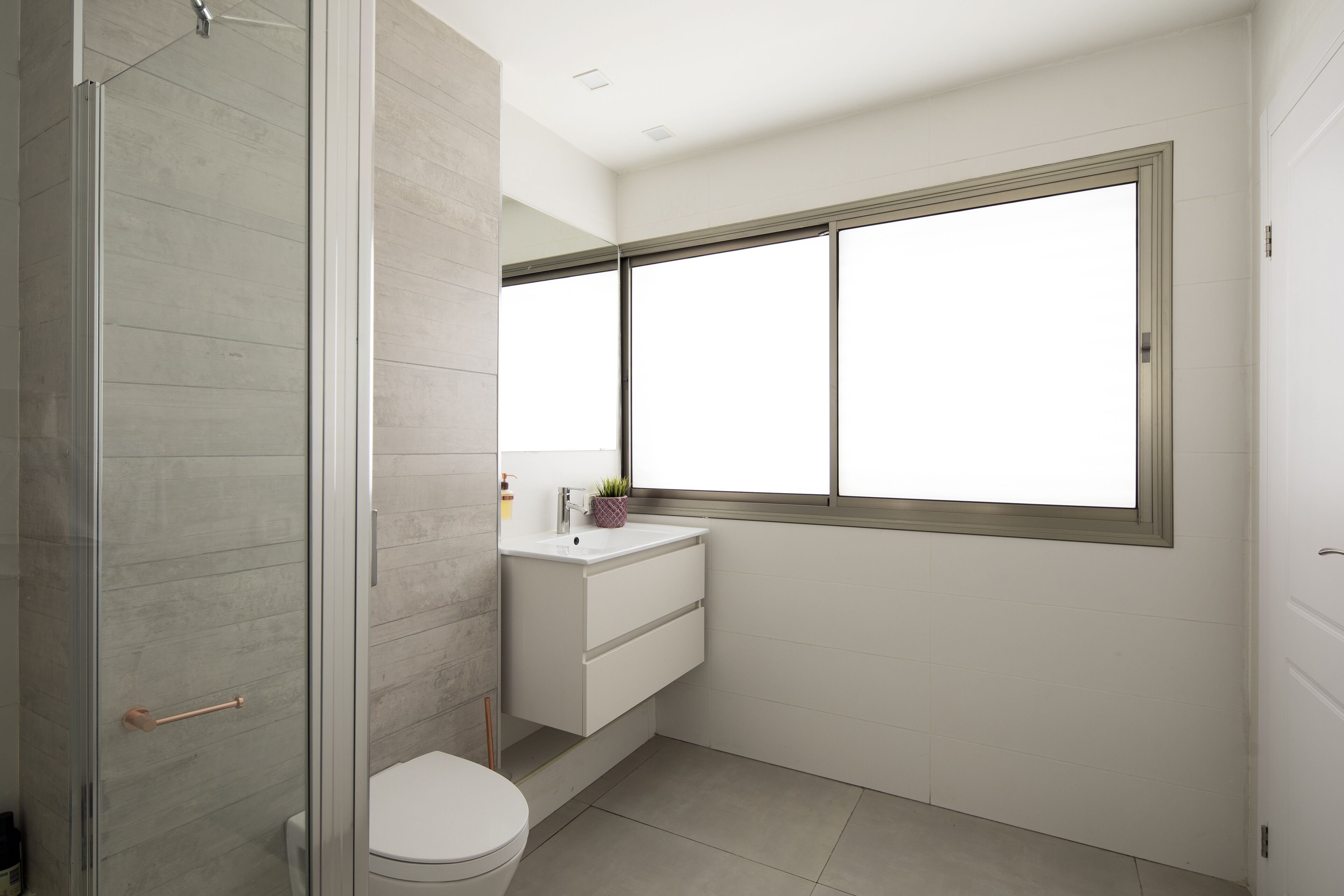 Un baño moderno con cabina de ducha de vidrio, inodoro blanco, lavabo con espejo sobre él y ventana que proporciona luz natural.