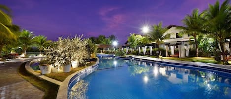Una piscina con borde de azulejos, rodeada de palmeras y un edificio con terraza cubierta.