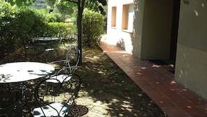 A courtyard with a table and chairs, surrounded by greenery and a building with a tiled pathway.