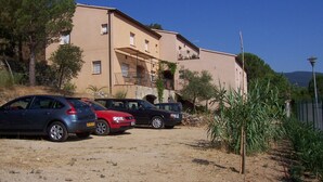 A building with a flat roof and a balcony, surrounded by parked cars and vegetation.
