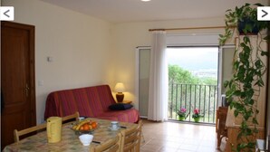 A cozy room with a sofa, a dining table with a fruit bowl, and a balcony with plants.