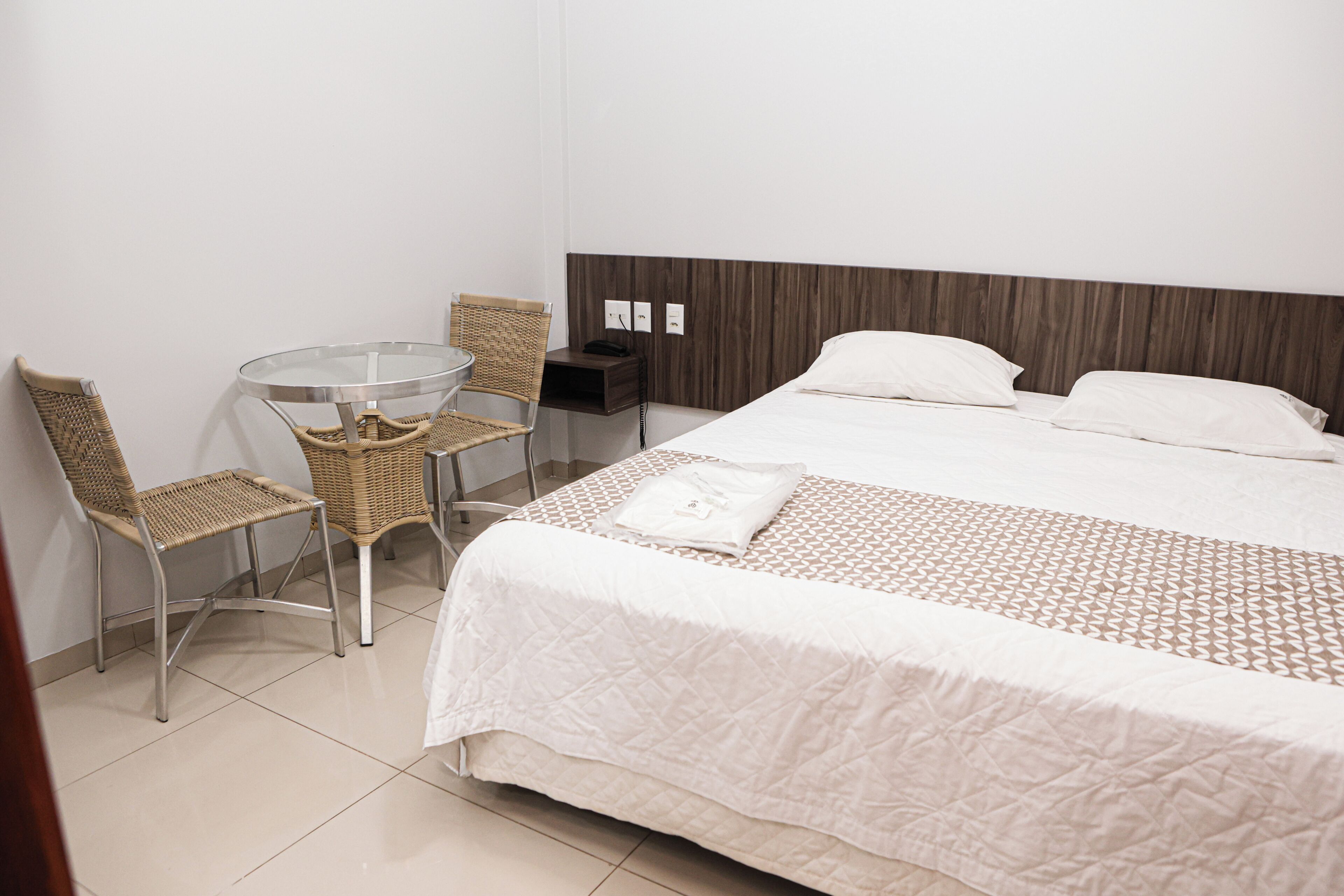 Une chambre d’hôtel comprenant un lit, deux fauteuils en osier, une table en verre, une table de chevet et une prise électrique murale.