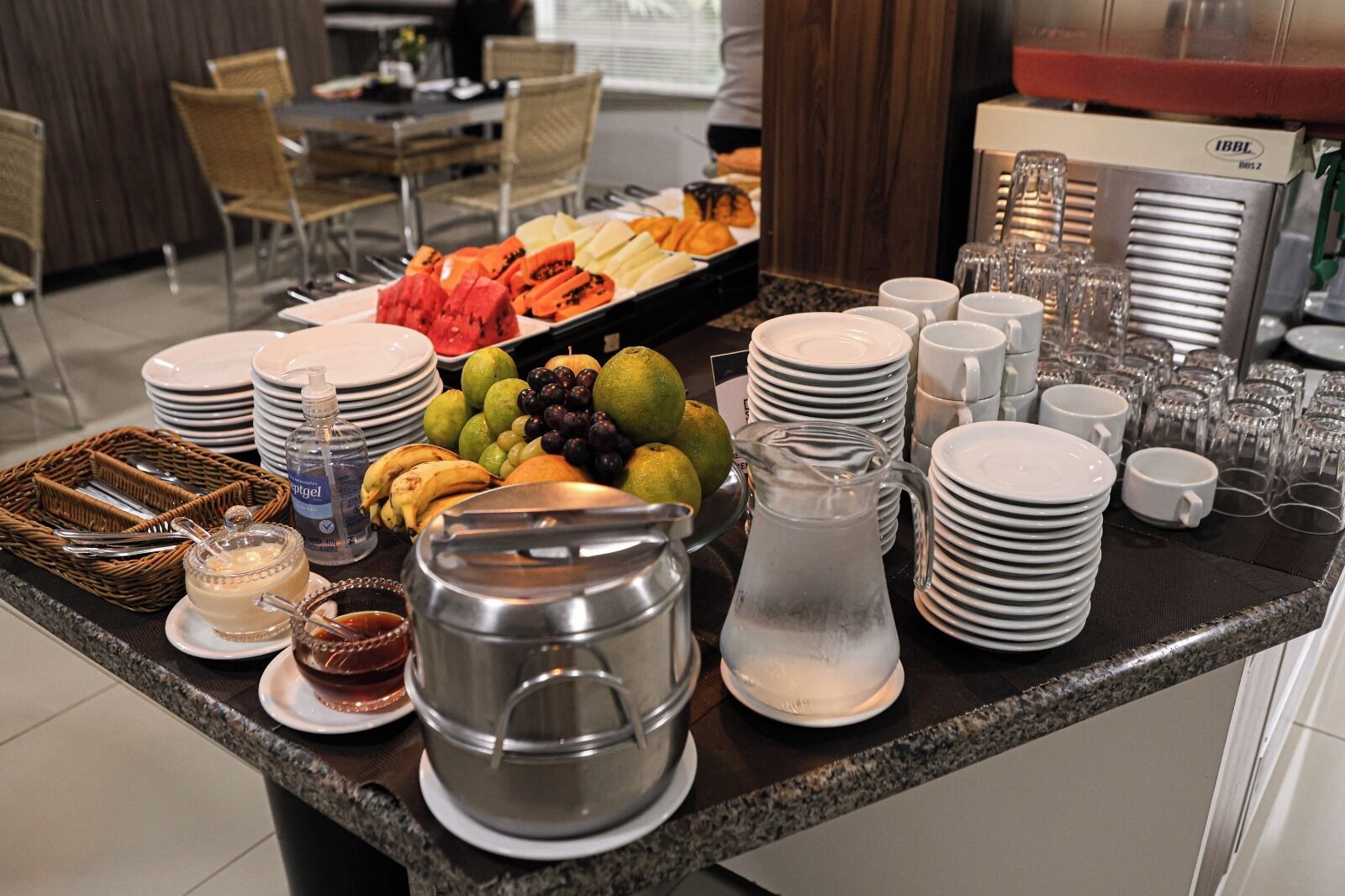 Een buffetopstelling met diverse soorten fruit, een kan en stapels borden en bekers.