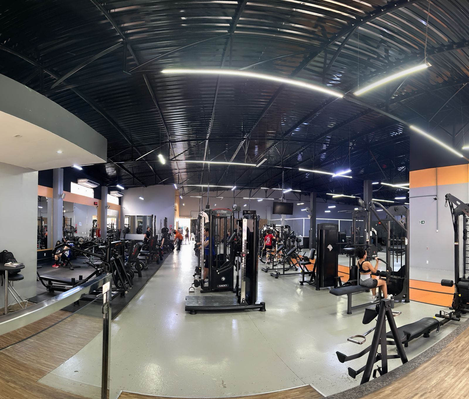 Un gimnasio con diversos equipos de ejercicio, un espejo grande en la pared y un techo alto con vigas a la vista.