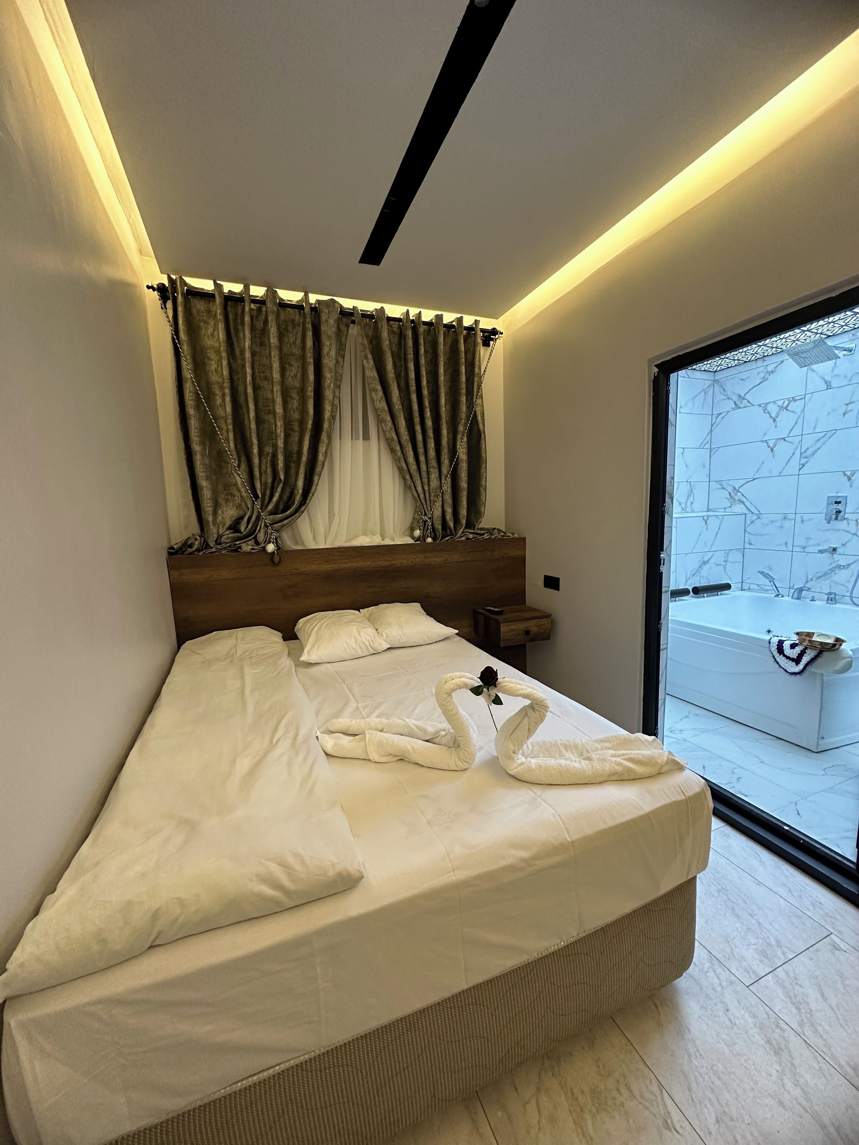 A modern bedroom with a bed, a nightstand, a window with curtains, and a view of a bathtub outside.