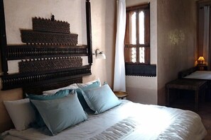 A bedroom with a large bed, multiple pillows, a wooden headboard, a small table, and a window with curtains. - Dar Blues (Ait Sedrate Jbel El Soufla)