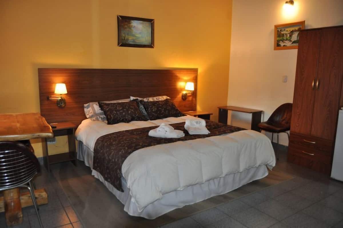 A hotel room with a large bed, two bedside lamps, a wooden headboard, a desk with a chair, and a wardrobe.