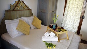 Una cama bien hecha con sábanas blancas, cabecera de madera con adornos de estrellas, una mesita de noche con toallas, una bandeja con frutas y un florero con flores.