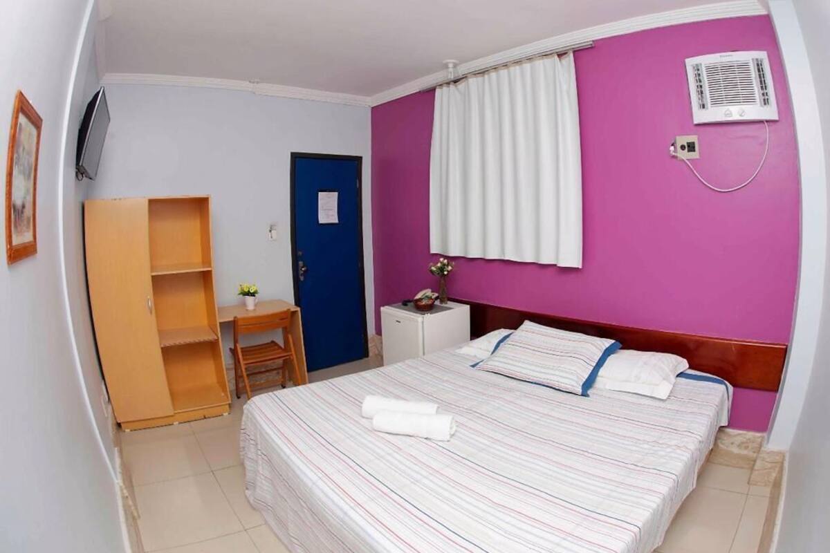 A hotel room with a bed, a desk, a wardrobe, a door, a window with curtains, and an air conditioning unit.