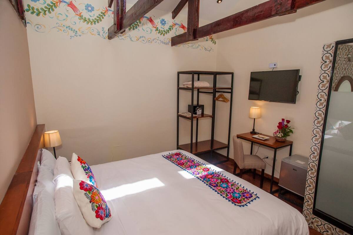 A bedroom with a bed, bedside lamps, a desk with a chair, a television, and a wardrobe.
