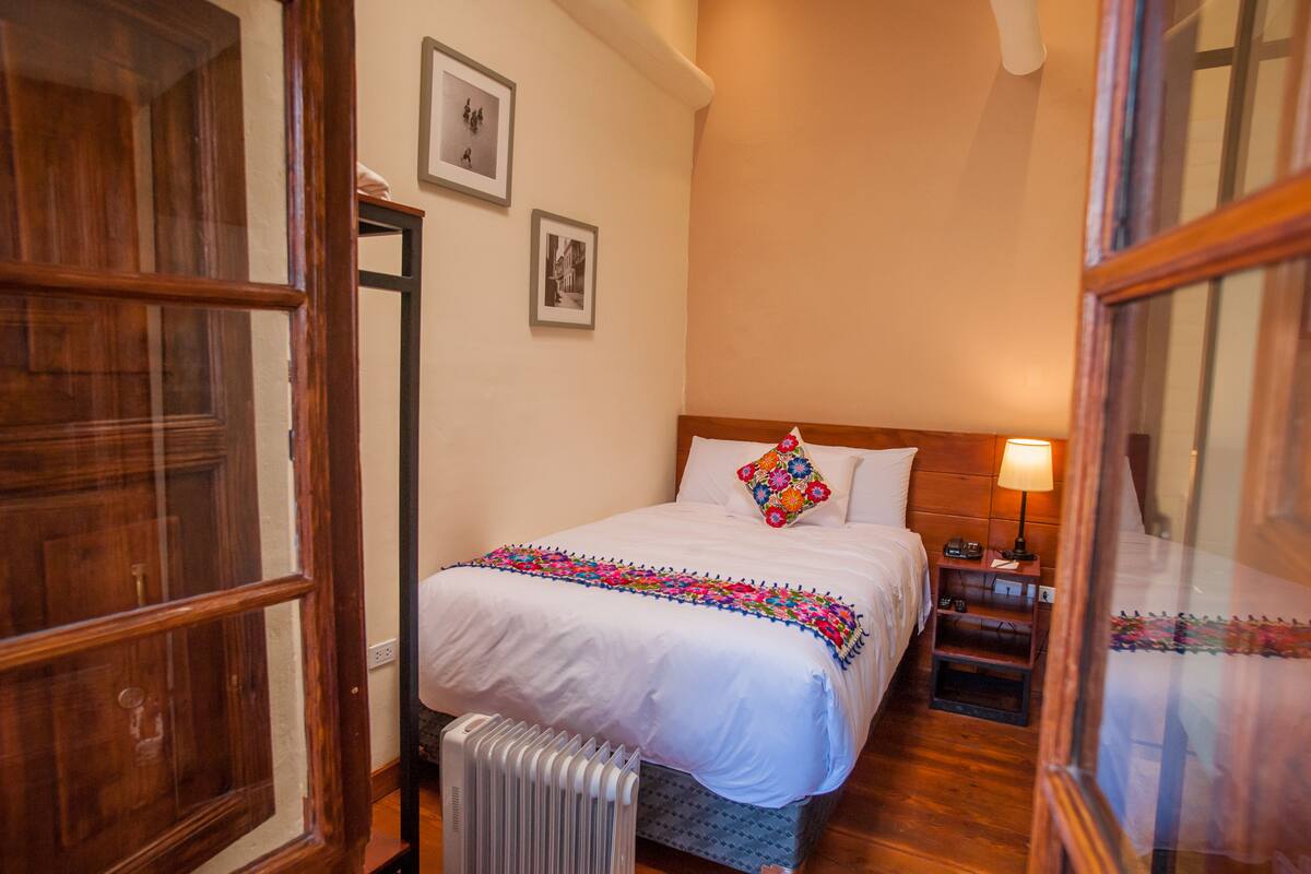 A bedroom with a wooden headboard, a white bedspread, a colorful pillow, a bedside table with a lamp, and a radiator.