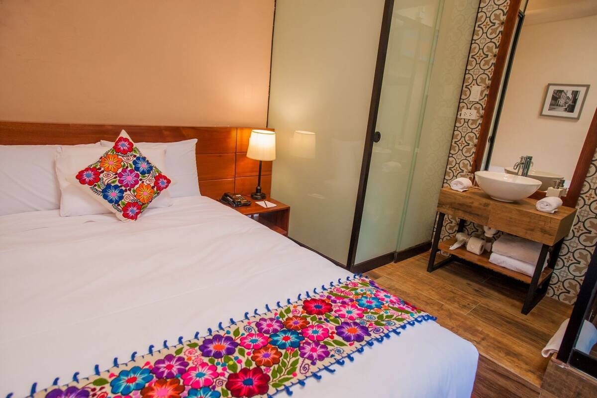 A hotel room with a bed, a nightstand, a lamp, a bathroom with a sink and mirror, and a patterned shower curtain.