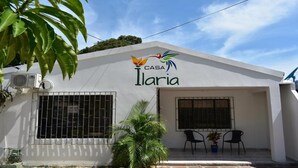 Un edificio blanco con un letrero que dice "CASA Ilaria" y un logotipo colorido. Hay dos ventanas con barrotes de metal, un pequeño espacio al aire libre con dos sillas y una mesa, y una planta en maceta.
