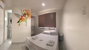 Un dormitorio con cama, ventana con persianas, aire acondicionado de pared y un baño a la vista en el fondo.