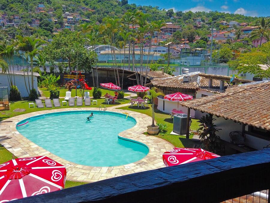 Una piscina con diseño en forma de corazón, rodeada de sillones de descanso y sombrillas, con vista a una zona residencial en la ladera.