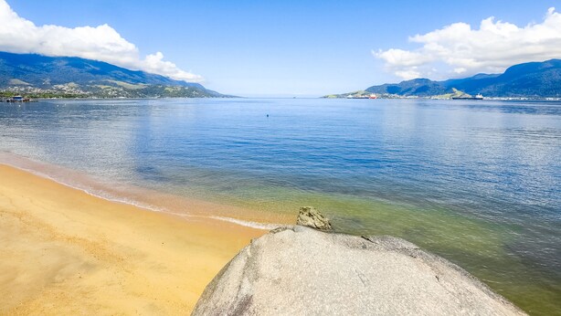 Una playa con agua cristalina, una roca grande en primer plano y montañas al fondo.