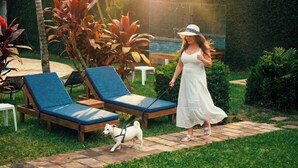Una mujer con un vestido blanco pasea un perro por un sendero con sillones de descanso y plantas tropicales.
