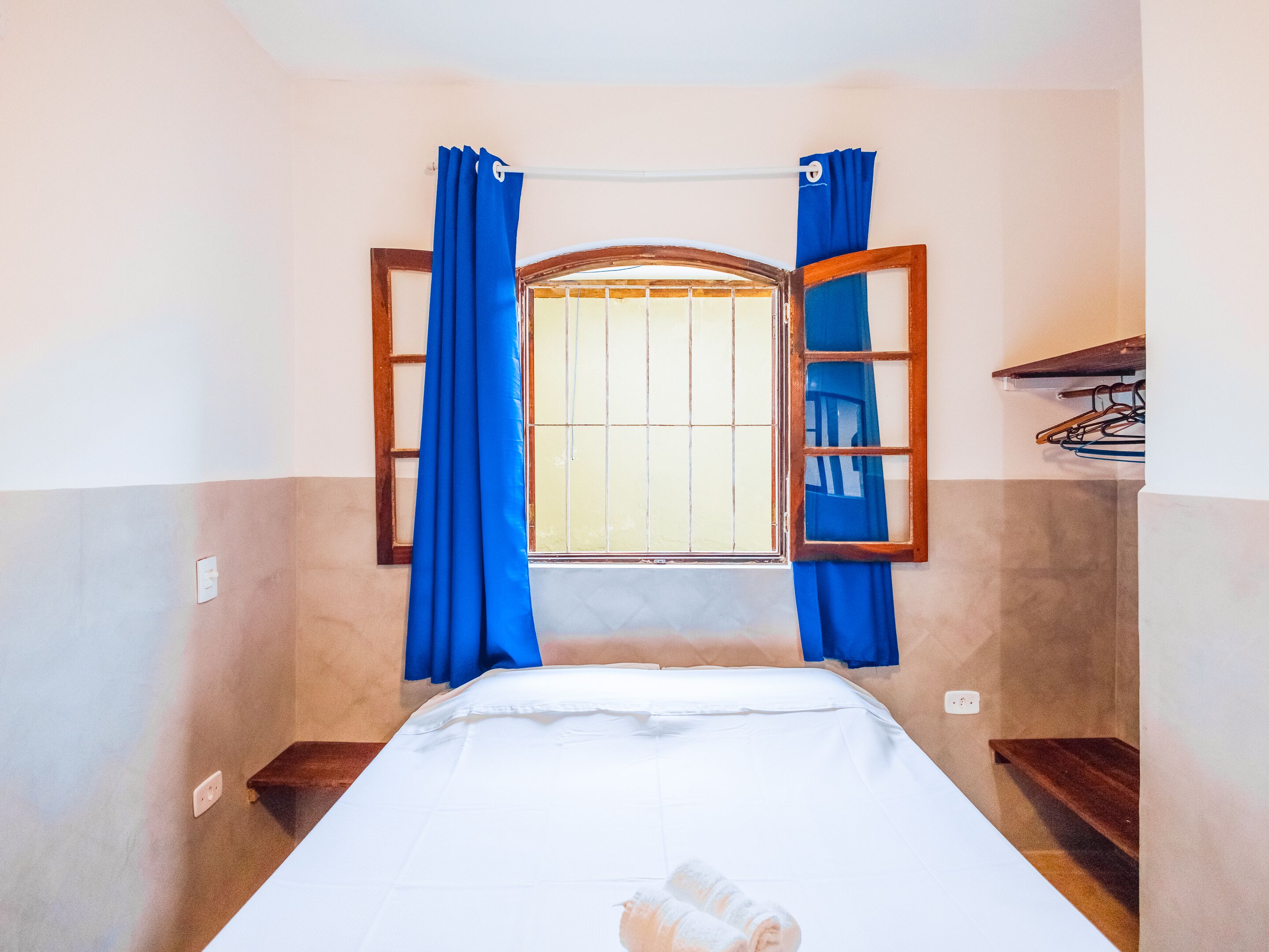 Habitación pequeña con una cama individual, ventana, cortinas azules y un estante de madera.