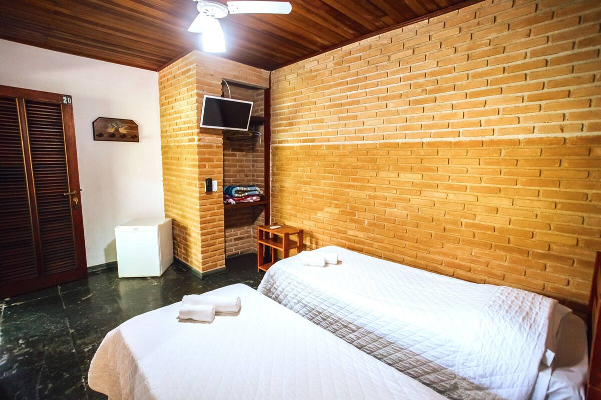Una habitación de hotel con pared de ladrillo, una cama con ropa blanca, un televisor montado en la pared y un ventilador de techo de madera.