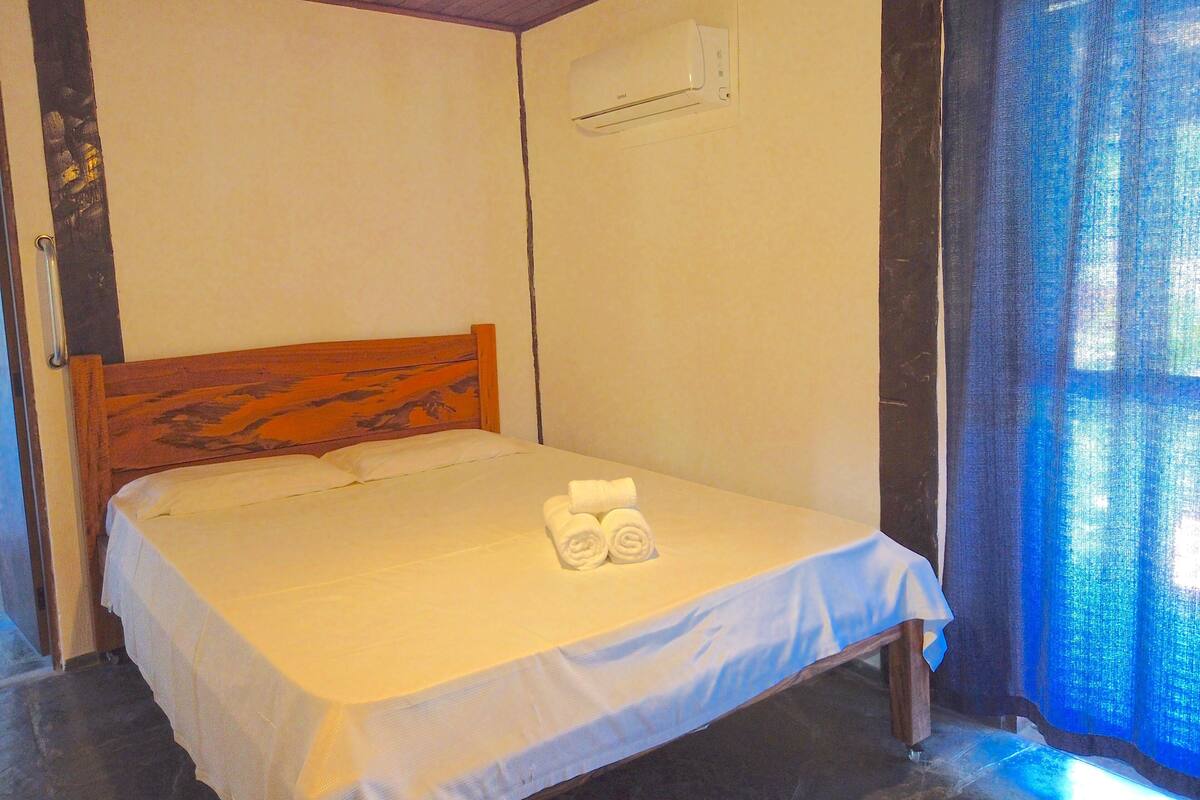 Una cama individual con cabecera de madera, una colcha blanca y dos toallas enrolladas. Un equipo de aire acondicionado está montado en la pared sobre la cama.