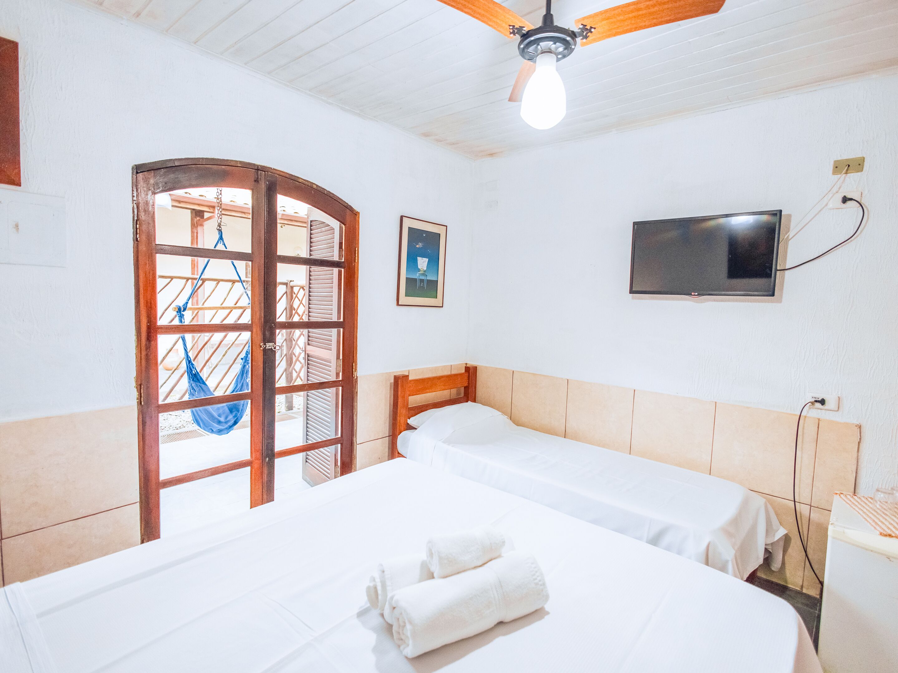 Quarto de hotel com duas camas, um ventilador de teto, televisão e porta que dá para área externa com rede.