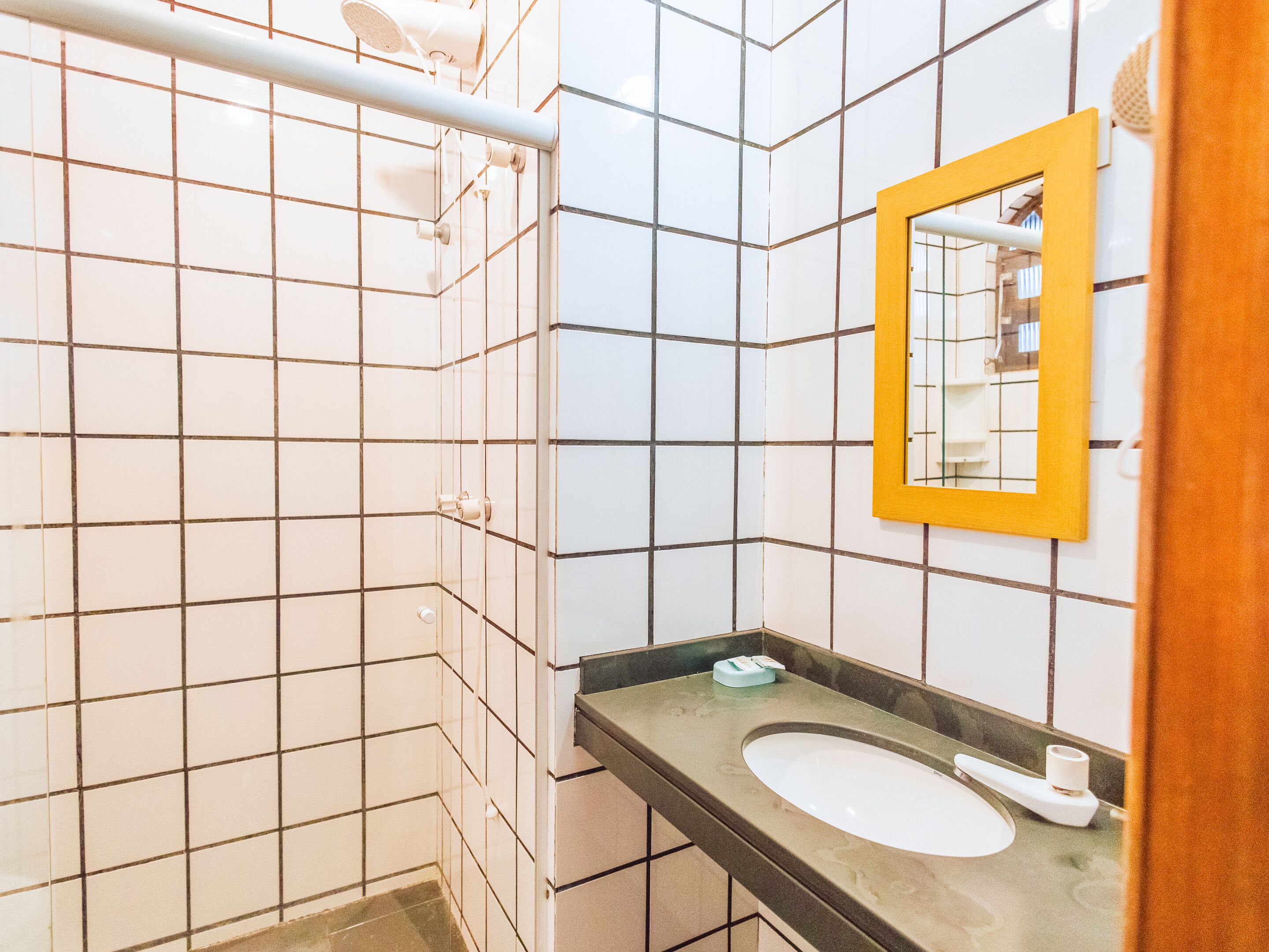 Un baño con paredes de azulejos blancos, un espejo con marco amarillo, un lavabo y una zona de ducha.
