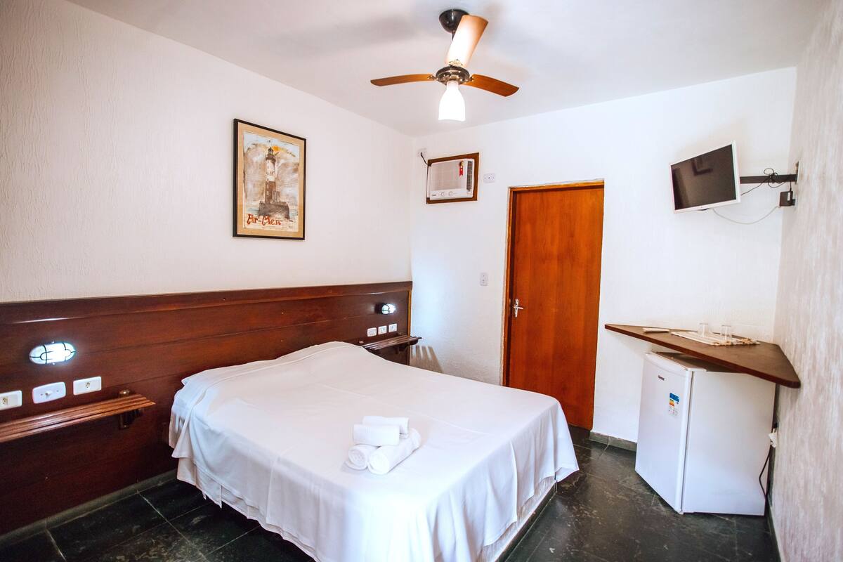 Habitación de hotel con cama, ventilador de techo, televisor montado en la pared, refrigerador pequeño y una puerta.