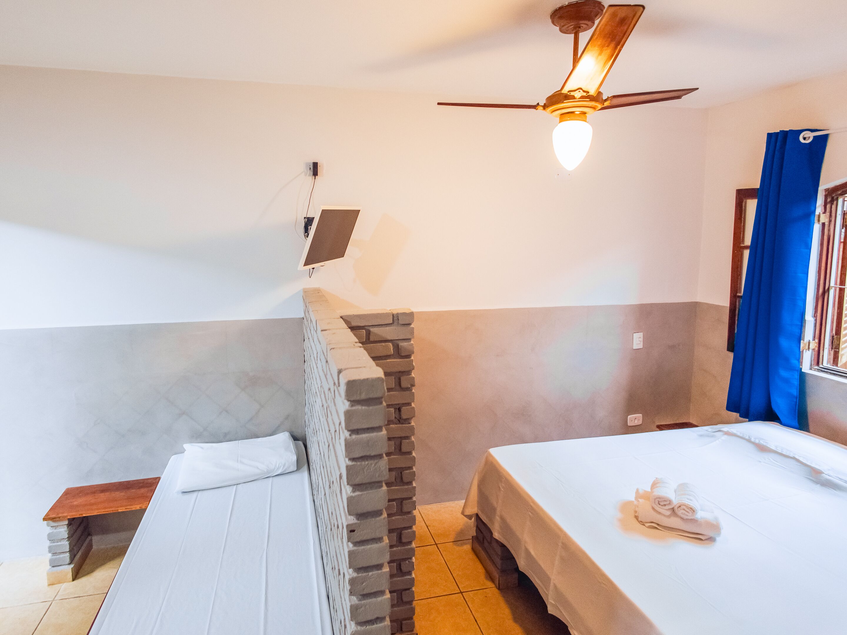 A bedroom with a bed, a small table, a brick wall, a ceiling fan, and a window with curtains.