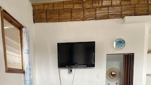 Una sala de estar con un sofá azul, un sillón verde azulado, una mesita blanca pequeña, un televisor montado en la pared y un refrigerador.